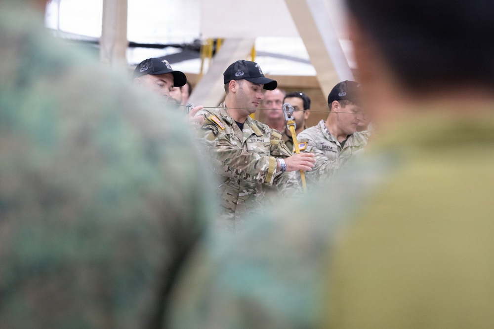 The image shows U.S. Army personnel in multicam/OCP uniforms conducting what appears to be a training demonstration or instruction session indoors. A central figure wearing a 'U.S. ARMY' labeled uniform is handling rope or rigging equipment, likely demonstrating a technique to an audience. The setting appears to be a training facility or hangar with wooden structural elements visible in the background.
