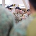 The image shows U.S. Army personnel in multicam/OCP uniforms conducting what appears to be a training demonstration or instruction session indoors. A central figure wearing a 'U.S. ARMY' labeled uniform is handling rope or rigging equipment, likely demonstrating a technique to an audience. The setting appears to be a training facility or hangar with wooden structural elements visible in the background.