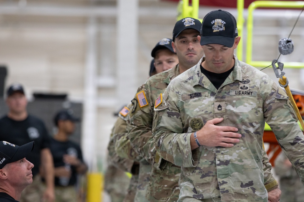The image shows U.S. Army soldiers in OCP (Operational Camouflage Pattern) uniforms at what appears to be an indoor military event or competition. The foreground soldier's name tape reads 'STONE' and displays U.S. Army insignia along with a Staff Sergeant rank patch. Several individuals in black caps and civilian-style clothing are also present in the background.