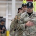The image shows U.S. Army soldiers in OCP (Operational Camouflage Pattern) uniforms at what appears to be an indoor military event or competition. The foreground soldier's name tape reads 'STONE' and displays U.S. Army insignia along with a Staff Sergeant rank patch. Several individuals in black caps and civilian-style clothing are also present in the background.