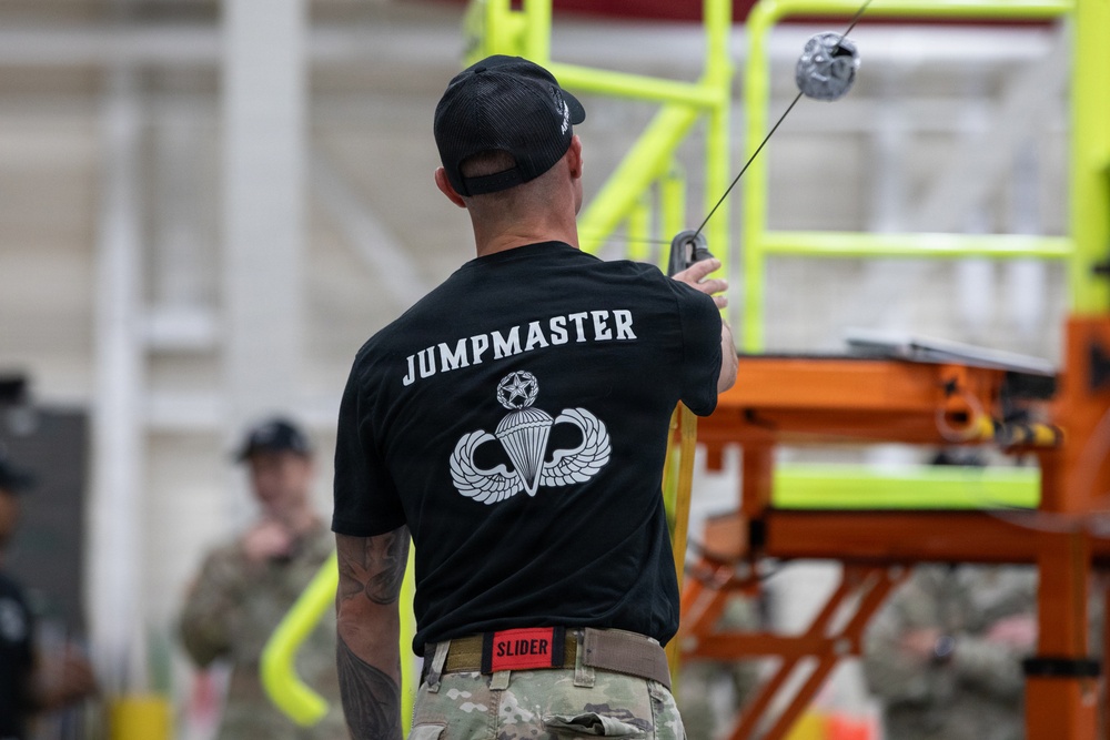 A military personnel member wearing a black 'JUMPMASTER' t-shirt with Master Parachutist wings insignia is shown from behind, holding what appears to be a static line or cable with a snap hook. The individual is wearing multicam/OCP pattern pants and a black baseball cap, standing in what appears to be an indoor training facility. Other uniformed personnel are visible in the background near yellow and orange equipment structures.