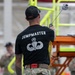 A military personnel member wearing a black 'JUMPMASTER' t-shirt with Master Parachutist wings insignia is shown from behind, holding what appears to be a static line or cable with a snap hook. The individual is wearing multicam/OCP pattern pants and a black baseball cap, standing in what appears to be an indoor training facility. Other uniformed personnel are visible in the background near yellow and orange equipment structures.