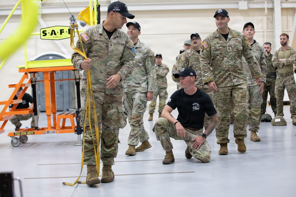 A group of U.S. Army soldiers in MultiCam/OCP camouflage uniforms are gathered inside what appears to be a large hangar or training facility. One soldier in the foreground is demonstrating a rigging or lifting technique using yellow straps, while another individual in a black t-shirt crouches nearby observing. The setting suggests a training exercise or equipment demonstration.