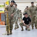 A group of U.S. Army soldiers in MultiCam/OCP camouflage uniforms are gathered inside what appears to be a large hangar or training facility. One soldier in the foreground is demonstrating a rigging or lifting technique using yellow straps, while another individual in a black t-shirt crouches nearby observing. The setting suggests a training exercise or equipment demonstration.