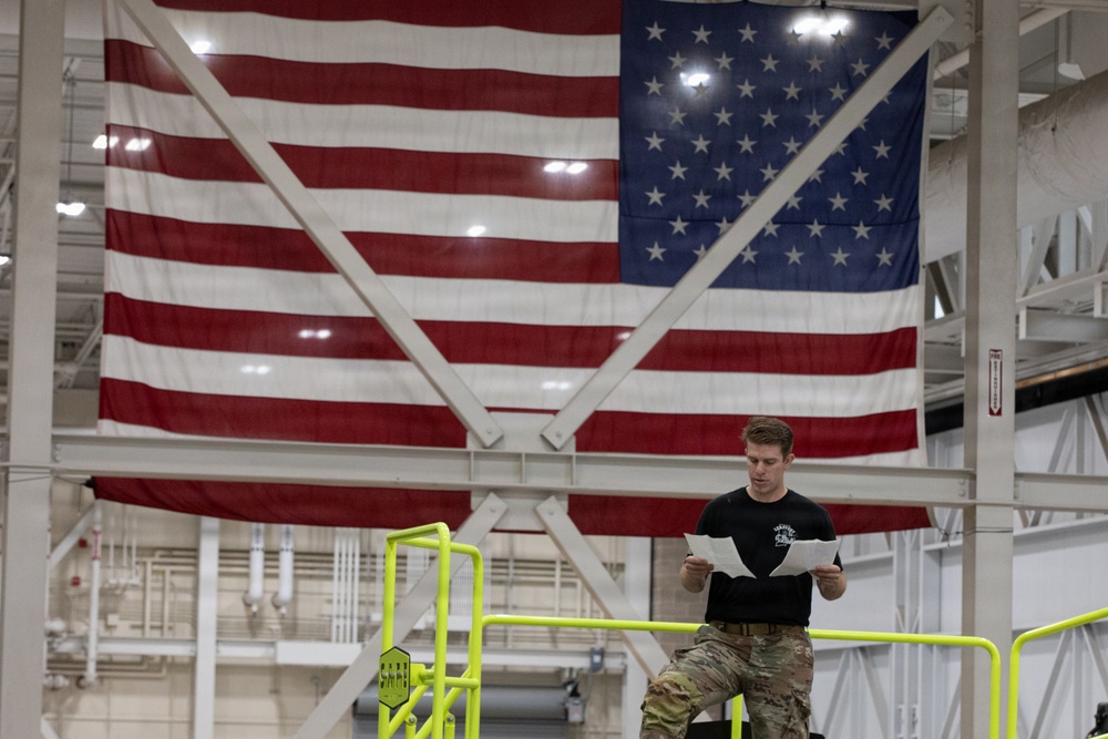 A military service member in camouflage pants and a black t-shirt is seated on an elevated platform with yellow safety railings, reading documents. Behind him, a large American flag is prominently displayed on the wall of what appears to be a large industrial or hangar facility. The setting suggests a military installation or aerospace facility.