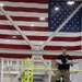 A military service member in camouflage pants and a black t-shirt is seated on an elevated platform with yellow safety railings, reading documents. Behind him, a large American flag is prominently displayed on the wall of what appears to be a large industrial or hangar facility. The setting suggests a military installation or aerospace facility.