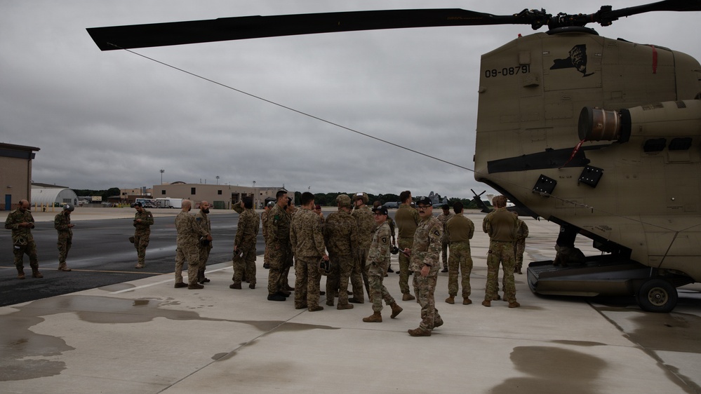 A group of military personnel in camouflage uniforms are gathered on a flight line tarmac next to a large Army CH-47 Chinook helicopter. The helicopter bears the tail number 09-08791 and displays a New York state insignia, indicating it belongs to the New York Army National Guard. The scene appears to be a briefing, pre-mission assembly, or post-landing gathering at a military airfield.
