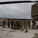 A group of military personnel in camouflage uniforms are gathered on a flight line tarmac next to a large Army CH-47 Chinook helicopter. The helicopter bears the tail number 09-08791 and displays a New York state insignia, indicating it belongs to the New York Army National Guard. The scene appears to be a briefing, pre-mission assembly, or post-landing gathering at a military airfield.