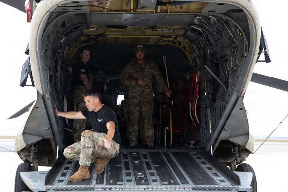 Three personnel are visible inside the open rear cargo bay of a large military helicopter, likely a CH-47 Chinook or similar heavy-lift rotary aircraft. One individual in a black t-shirt and camouflage pants is crouched at the edge of the lowered ramp, while two others stand inside the cargo hold. The scene appears to depict a pre-flight check, loading operation, or inspection on a tarmac.