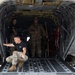 Three personnel are visible inside the open rear cargo bay of a large military helicopter, likely a CH-47 Chinook or similar heavy-lift rotary aircraft. One individual in a black t-shirt and camouflage pants is crouched at the edge of the lowered ramp, while two others stand inside the cargo hold. The scene appears to depict a pre-flight check, loading operation, or inspection on a tarmac.