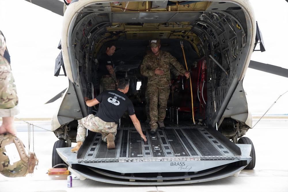 Military personnel are working around the open rear cargo ramp of a large military helicopter, likely a CH-47 Chinook. Several individuals in military uniforms and civilian-style clothing are loading, inspecting, or preparing the aircraft. The text 'BRASS MONKEY' is visible on the rear ramp edge.