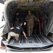 Military personnel are working around the open rear cargo ramp of a large military helicopter, likely a CH-47 Chinook. Several individuals in military uniforms and civilian-style clothing are loading, inspecting, or preparing the aircraft. The text 'BRASS MONKEY' is visible on the rear ramp edge.