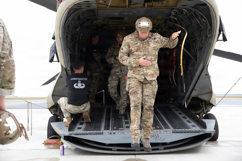 Military personnel are exiting or conducting operations at the rear ramp of a large military helicopter, likely a CH-47 Chinook. One soldier in multicam uniform and tactical helmet is stepping off the ramp while gesturing, and another individual wearing a black 'JUMPMASTER' t-shirt is crouching near the ramp. The scene appears to take place on a tarmac or airfield.