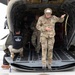 Military personnel are exiting or conducting operations at the rear ramp of a large military helicopter, likely a CH-47 Chinook. One soldier in multicam uniform and tactical helmet is stepping off the ramp while gesturing, and another individual wearing a black 'JUMPMASTER' t-shirt is crouching near the ramp. The scene appears to take place on a tarmac or airfield.