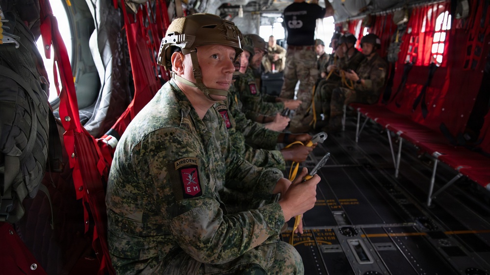 Military personnel in full combat gear are seated inside what appears to be a military transport aircraft, likely a C-130 Hercules, preparing for an airborne operation. The foreground soldier wears a digital camouflage uniform with an 'AIRBORNE' tab and a unit patch, along with a tactical helmet. Multiple soldiers are seated along the red cargo netting seats of the aircraft interior.