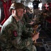 Military personnel in full combat gear are seated inside what appears to be a military transport aircraft, likely a C-130 Hercules, preparing for an airborne operation. The foreground soldier wears a digital camouflage uniform with an 'AIRBORNE' tab and a unit patch, along with a tactical helmet. Multiple soldiers are seated along the red cargo netting seats of the aircraft interior.