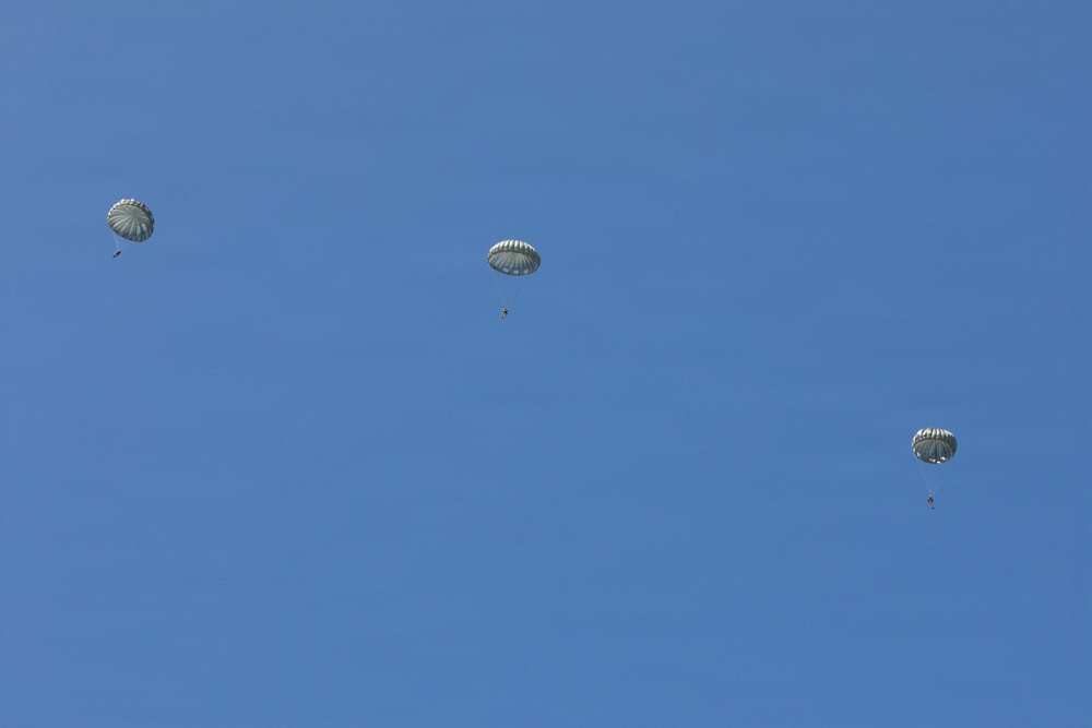 Three military-style parachutes are visible against a clear blue sky, each with a suspended figure beneath them. The parachutes appear to be round canopy designs consistent with military airborne operations. The image was taken from the ground looking upward.