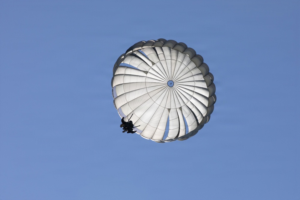 A white round parachute is fully deployed against a clear blue sky, with a person suspended beneath it in silhouette. The parachute appears to be a military-style round canopy design, photographed from below at an angle. The image captures a descent in progress under calm, clear weather conditions.