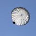 A white round parachute is fully deployed against a clear blue sky, with a person suspended beneath it in silhouette. The parachute appears to be a military-style round canopy design, photographed from below at an angle. The image captures a descent in progress under calm, clear weather conditions.