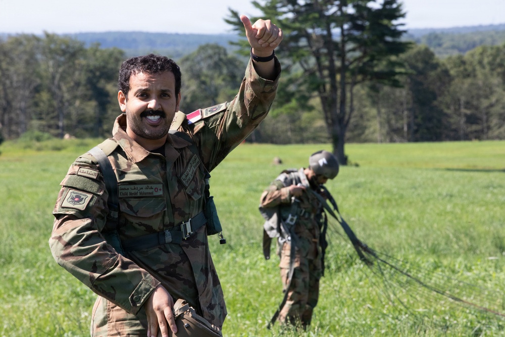 A smiling soldier in camouflage uniform gives a thumbs-up gesture in an open grassy field, appearing to celebrate or signal success after what appears to be a parachute landing. Name tape on the uniform reads 'Khalid Marif Mohammed' with Arabic script and a flag patch visible. A second soldier is visible in the background gathering parachute lines.