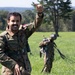 A smiling soldier in camouflage uniform gives a thumbs-up gesture in an open grassy field, appearing to celebrate or signal success after what appears to be a parachute landing. Name tape on the uniform reads 'Khalid Marif Mohammed' with Arabic script and a flag patch visible. A second soldier is visible in the background gathering parachute lines.
