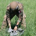 A soldier wearing camouflage uniform and a tan/olive helmet is kneeling in a grassy field, handling what appears to be a gray parachute or equipment bag. The soldier's uniform displays a Qatar national flag patch on the right shoulder, indicating affiliation with the Qatari armed forces. The setting appears to be an outdoor training or exercise environment with lush green grass.