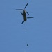 A tandem-rotor military helicopter, consistent with a CH-47 Chinook, is photographed from below against a clear blue sky. The helicopter appears to be conducting a sling load operation, with a suspended load hanging below it on a long line. The image was taken from a ground-level perspective looking upward.