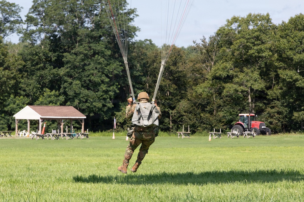 A military paratrooper is captured mid-landing on a grassy field, with parachute lines still taut above them. The soldier is wearing full combat gear including camouflage uniform, helmet, and tactical vest. The landing zone appears to be a rural field with a pavilion structure and a red tractor visible in the background.