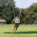 A military paratrooper is captured mid-landing on a grassy field, with parachute lines still taut above them. The soldier is wearing full combat gear including camouflage uniform, helmet, and tactical vest. The landing zone appears to be a rural field with a pavilion structure and a red tractor visible in the background.