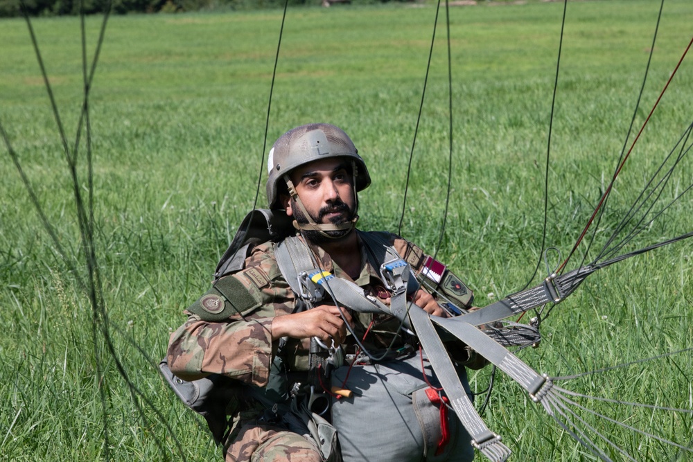 A military paratrooper is seated on a grassy field after a landing, still entangled in parachute suspension lines and harness straps. He is wearing camouflage uniform and a ballistic helmet, looking alert to his surroundings. The image captures the immediate post-landing phase of a parachute operation.