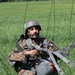 A military paratrooper is seated on a grassy field after a landing, still entangled in parachute suspension lines and harness straps. He is wearing camouflage uniform and a ballistic helmet, looking alert to his surroundings. The image captures the immediate post-landing phase of a parachute operation.