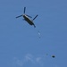 A military tandem-rotor helicopter, identifiable as a CH-47 Chinook, is captured in flight against a clear blue sky. The helicopter appears to be conducting an aerial delivery or sling-load operation, with objects visible below it on a line or cable. A parachute-deployed load is visible further below, suggesting an airdrop operation in progress.