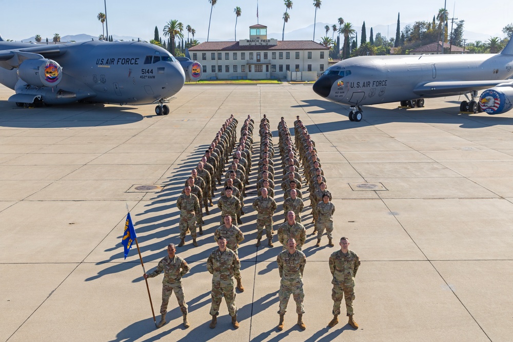 A large formation of U.S. Air Force personnel stands at attention on a flight line tarmac between two military aircraft. The formation is photographed from an elevated angle at March Field, with a historic Spanish Colonial Revival-style building visible in the background. A guidon bearer holds a blue unit flag at the front of the formation.
