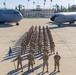 A large formation of U.S. Air Force personnel stands at attention on a flight line tarmac between two military aircraft. The formation is photographed from an elevated angle at March Field, with a historic Spanish Colonial Revival-style building visible in the background. A guidon bearer holds a blue unit flag at the front of the formation.