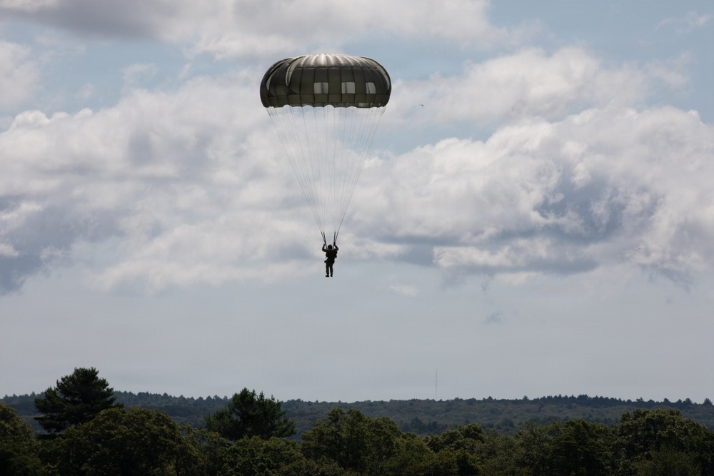 A single parachutist is descending under a dark olive/military-green round parachute canopy against a partly cloudy sky. The figure appears as a silhouette suspended beneath the fully deployed chute, with a forested landscape visible below. The scene suggests a military airborne operation or training exercise.