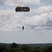 A single parachutist is descending under a dark olive/military-green round parachute canopy against a partly cloudy sky. The figure appears as a silhouette suspended beneath the fully deployed chute, with a forested landscape visible below. The scene suggests a military airborne operation or training exercise.