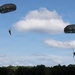 Two military paratroopers are descending under dark olive/green round parachutes against a partly cloudy blue sky. The parachutes appear to be standard military static-line round canopies, consistent with airborne operations. A treeline is visible at the bottom of the image, suggesting the jumpers are at a relatively low altitude near landing.