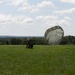 A military paratrooper has just landed in an open green field and is managing a deployed olive-drab parachute that is still partially inflated on the ground. The scene appears to be a training or operational jump exercise taking place in a rural area with tree lines visible on the horizon. The parachute canopy is billowing sideways, likely being dragged by wind, as the soldier works to collapse and control it.