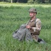 A male soldier in MultiCam/MTP camouflage uniform is kneeling in a grassy field while gathering or packing an olive-drab fabric item, likely a parachute or equipment bag. He is wearing a tactical helmet with rail attachments, a plate carrier or chest rig, and a black wristwatch. A small flag patch is visible on his right shoulder, consistent with a Union Jack, suggesting British military affiliation.