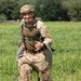 A smiling soldier in full military gear is standing in an open grassy field, appearing to have just landed or completed a parachute jump. He is wearing British MTP (Multi-Terrain Pattern) camouflage uniform with a tactical helmet and parachute harness. The background shows a rural landscape with dense tree line, a small structure, and farm equipment.