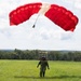 A parachutist has just landed or is in the final moments of landing on a green grassy field. The jumper is wearing military-style camouflage gear and a helmet, with a large red and white ram-air parachute canopy still inflated above them. The setting appears to be a rural drop zone surrounded by dense tree lines under a partly cloudy sky.