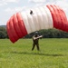 A parachutist has just landed on a green grassy field, still holding the control toggles of a fully inflated red and white ram-air parachute canopy. The individual is wearing camouflage military-style clothing and a blue helmet. The background shows a tree line and partly cloudy sky, suggesting a warm, clear day.