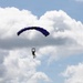 A single parachutist is descending through a partly cloudy sky using a ram-air (rectangular) parachute canopy in dark blue/purple coloring. The jumper appears to be holding control toggles and is suspended beneath the fully inflated canopy by visible suspension lines. The image is taken from a ground-level perspective looking upward.