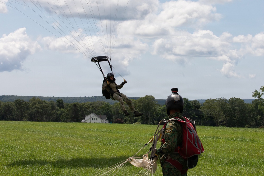 Two individuals in military-style uniforms and helmets are engaged in a parachuting activity on a green open field. One person is airborne and about to land, suspended beneath a small dark canopy parachute, while the second person stands on the ground with a deployed parachute at their feet. The setting appears to be a rural drop zone surrounded by trees and rolling hills under a partly cloudy sky.