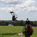 Two individuals in military-style uniforms and helmets are engaged in a parachuting activity on a green open field. One person is airborne and about to land, suspended beneath a small dark canopy parachute, while the second person stands on the ground with a deployed parachute at their feet. The setting appears to be a rural drop zone surrounded by trees and rolling hills under a partly cloudy sky.