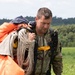 A male soldier in camouflage uniform is carrying a deployed parachute after landing in an open grassy field. He is gathering and holding the parachute canopy and suspension lines over his shoulder. The background shows a treeline and overcast sky consistent with a European landscape.