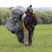 A parachutist is walking across a grassy field after landing, carrying a large bundled parachute canopy. The individual is wearing a full-face helmet with a camera mounted on top, a maroon t-shirt reading 'Parachute Instructor', tactical harness, and camouflage pants. The setting appears to be a rural drop zone surrounded by dense green trees under an overcast sky.