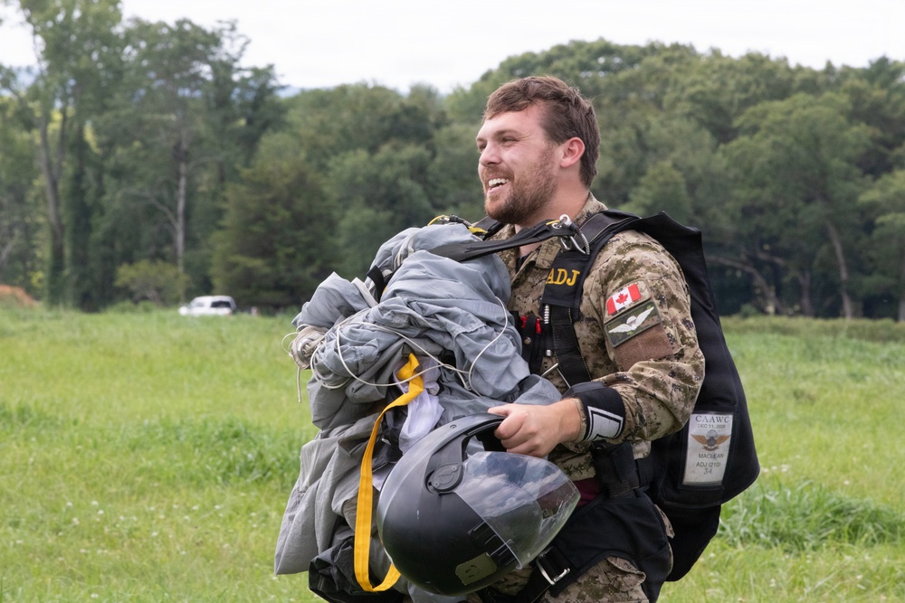 A smiling male military personnel, identified by a Canadian flag patch on his uniform, is carrying a bundled parachute and helmet after an apparent jump. He is wearing a camouflage uniform with tactical vest and appears to be on a grassy landing zone. His expression suggests a successful and enjoyable parachute operation.