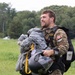 A smiling male military personnel, identified by a Canadian flag patch on his uniform, is carrying a bundled parachute and helmet after an apparent jump. He is wearing a camouflage uniform with tactical vest and appears to be on a grassy landing zone. His expression suggests a successful and enjoyable parachute operation.
