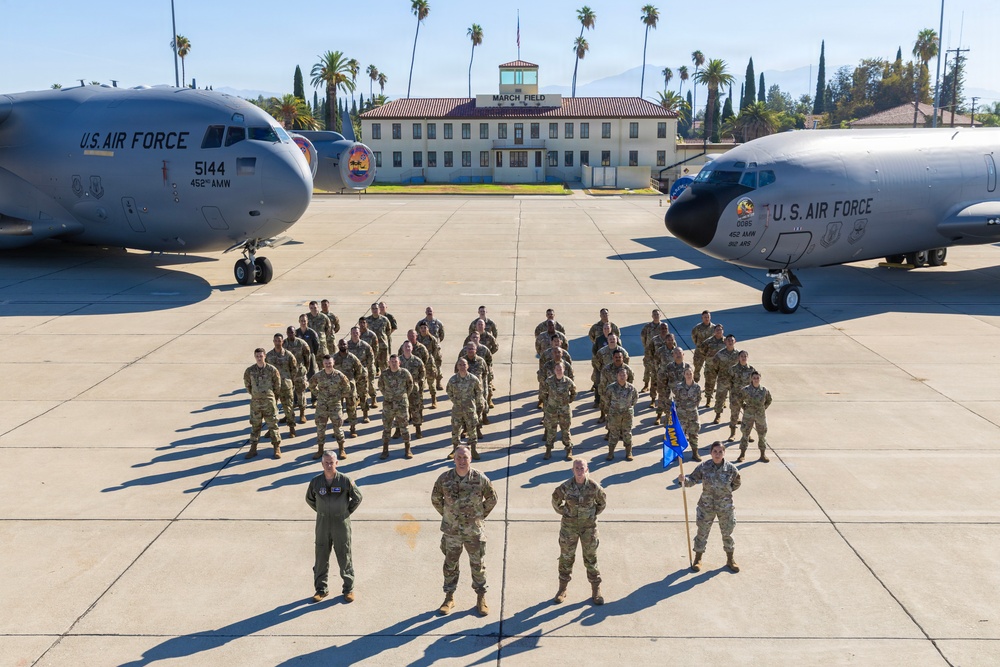 A formal group photograph of U.S. Air Force personnel standing in formation on a flight line tarmac at March Field Air Reserve Base. Two C-130 Hercules aircraft bearing '452 AMW' (452nd Air Mobility Wing) markings flank the group on either side. One service member holds a blue unit guidon flag, and a historic building labeled 'March Field' is visible in the background.
